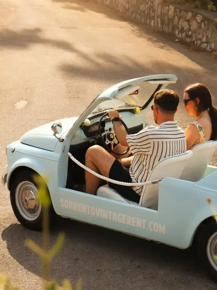 Couple driving a sky blue Fiat 500 Jolly along a shaded coastal road near Sorrento