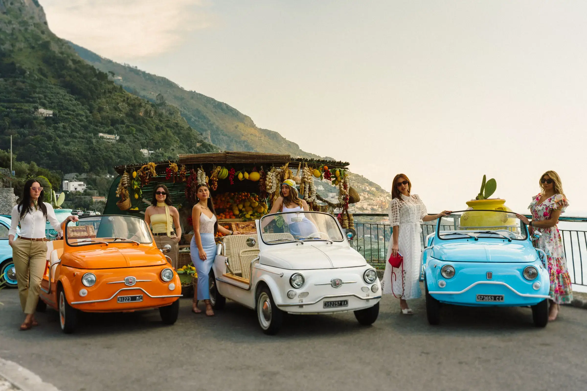 Three vintage Fiat 500 Jolly — orange, white and sky blue — lined up by a fruit stall overlooking the Amalfi Coast