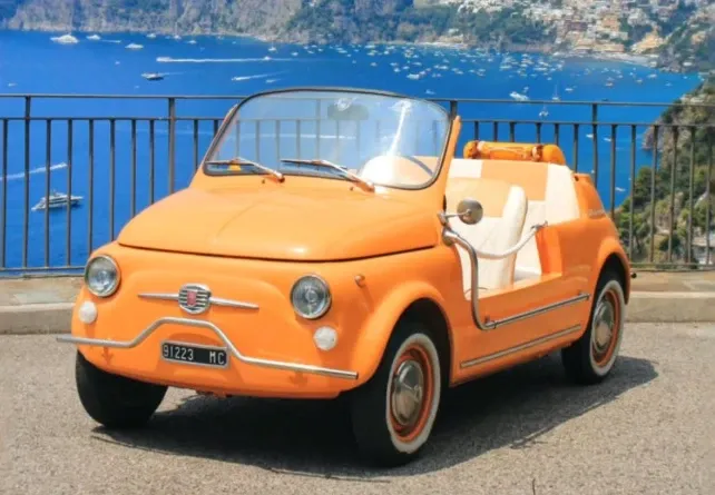 Orange Fiat 500 Jolly with wicker seats parked above the bay, boats moored in the water below