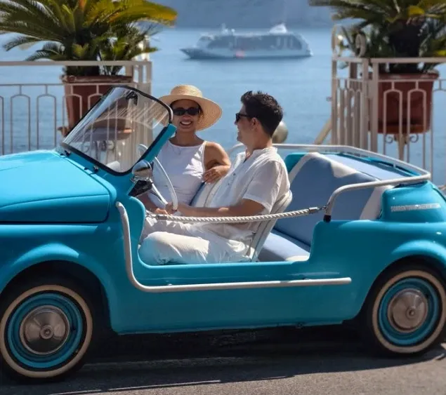 Couple in white riding a sky blue Fiat 500 Jolly along the seafront, a cruise ship on the horizon