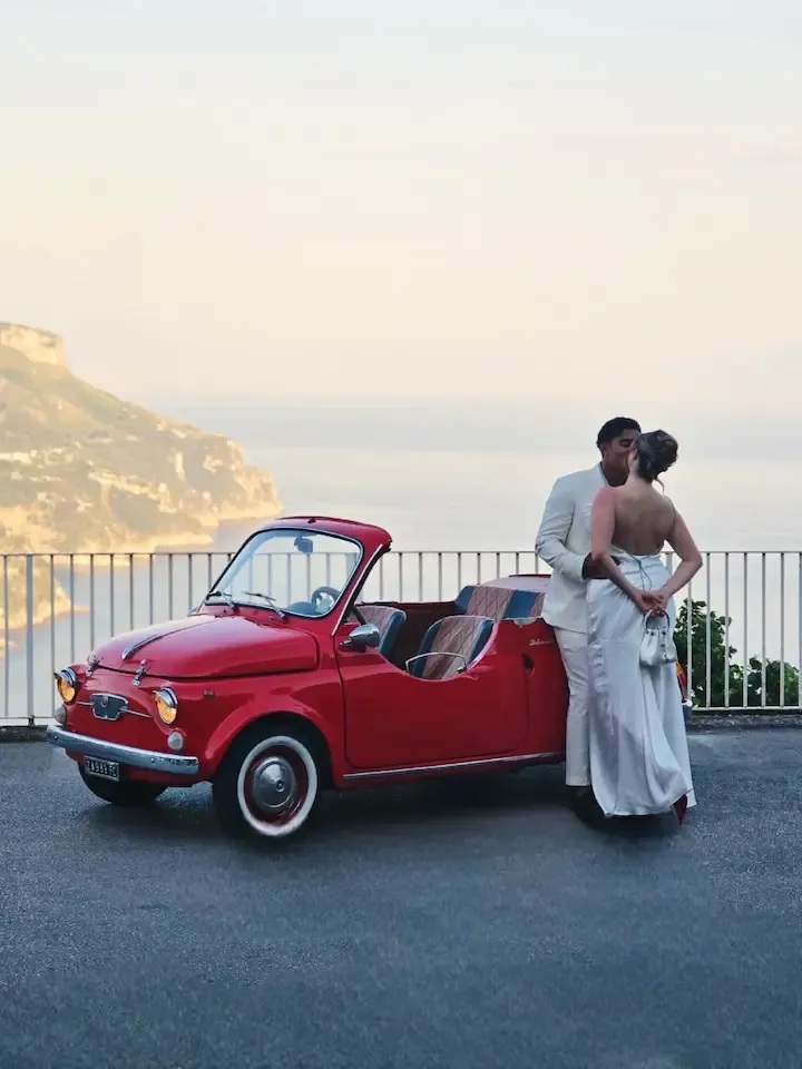 Bride and groom embracing beside a red vintage Fiat 500 Jolly above the sea at sunset